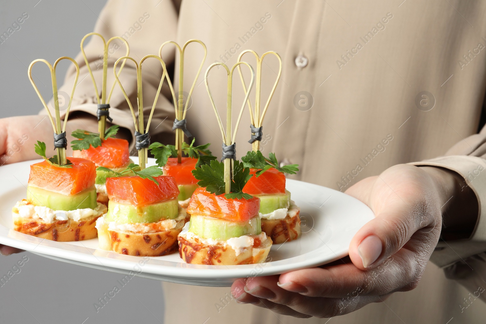 Photo of Woman holding tasty canapes with salmon, cucumber, bread and cream cheese on grey background, closeup