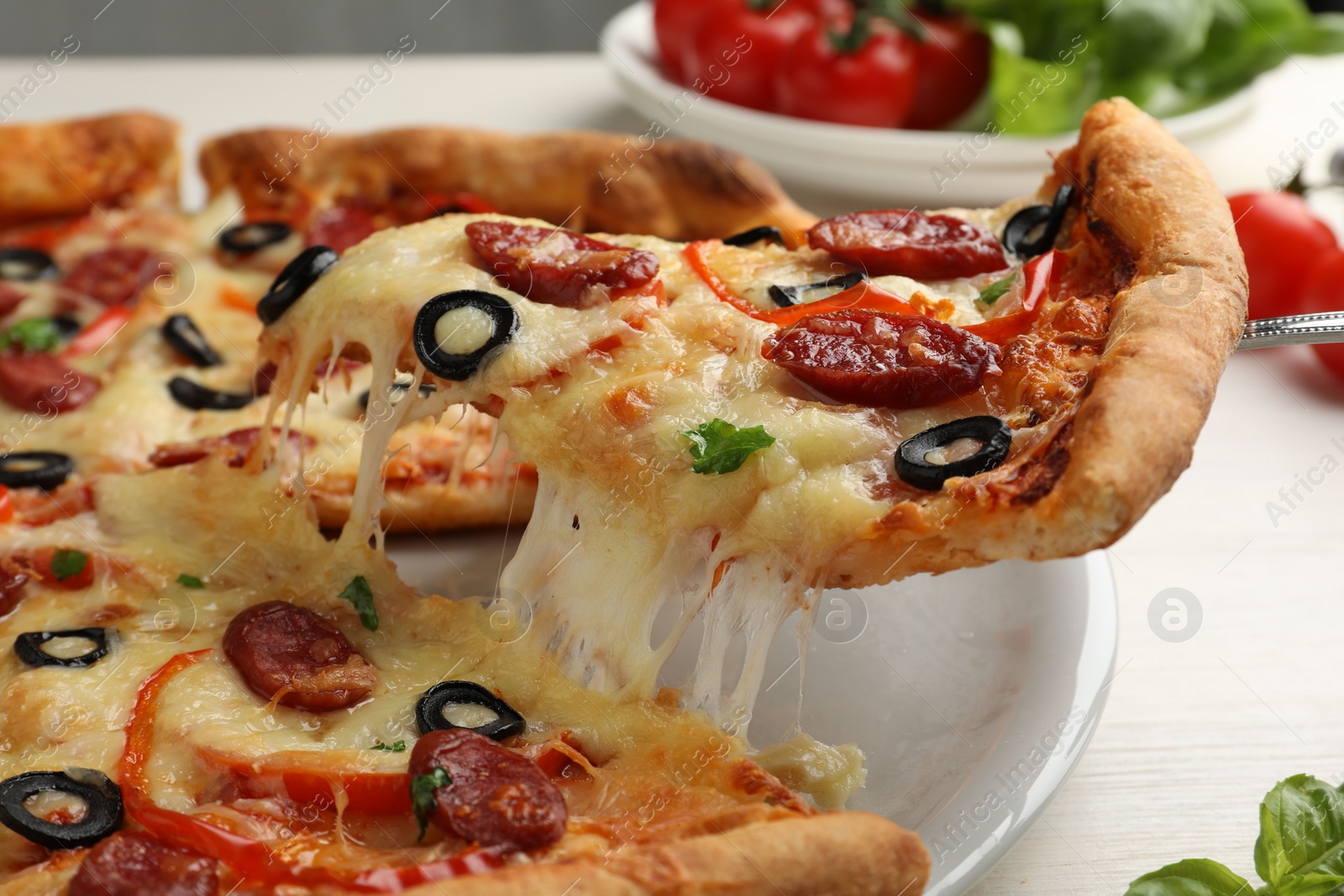 Photo of Taking piece of tasty pizza with smoked sausages, olives, pepper and parsley at light wooden table, closeup