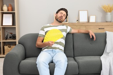 Photo of Bearded man waving yellow hand fan to cool himself on sofa at home