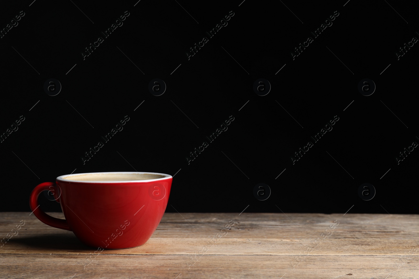 Photo of Red ceramic cup with hot aromatic coffee on wooden table against black background, space for text