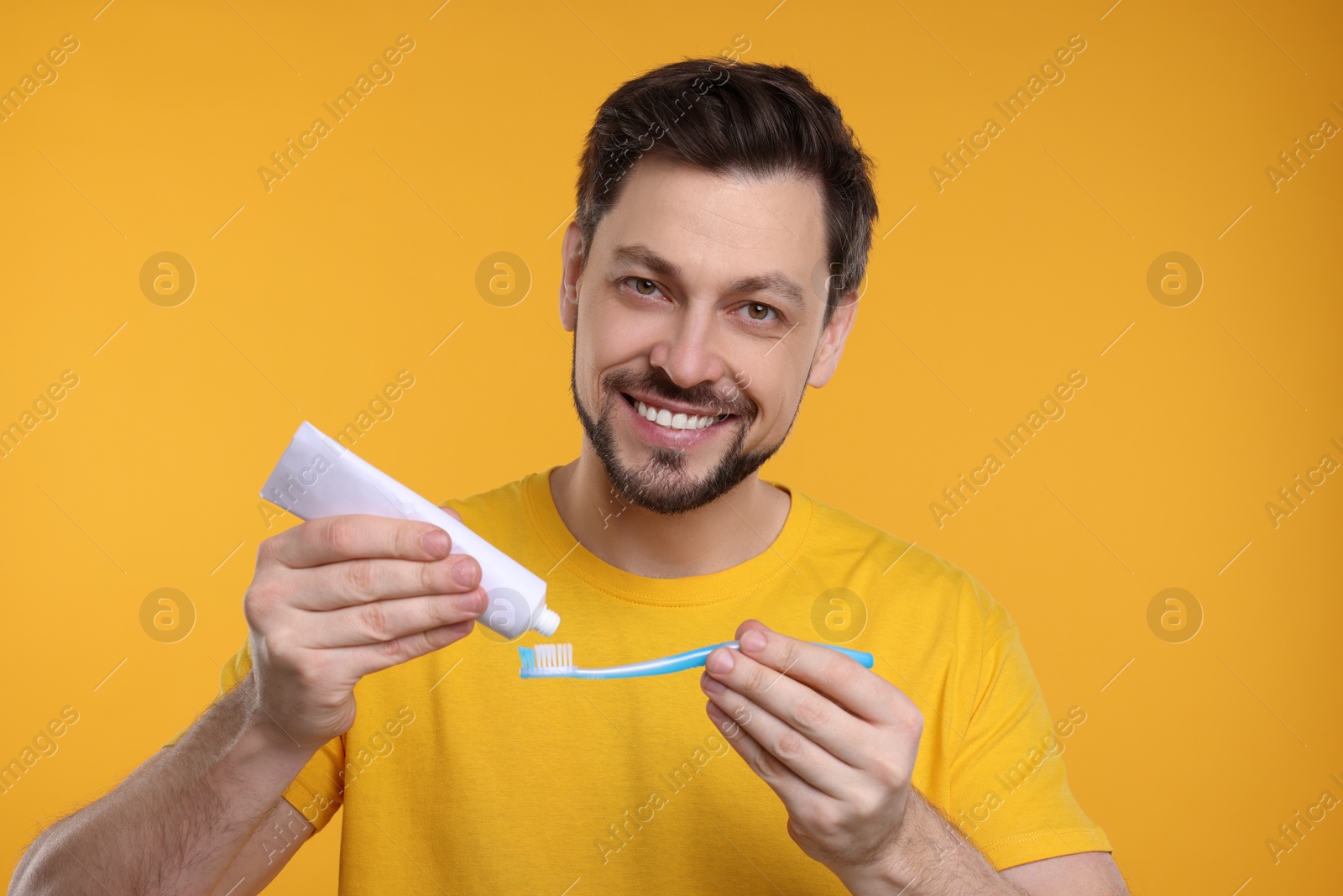 Photo of Happy man squeezing toothpaste from tube onto plastic toothbrush on yellow background