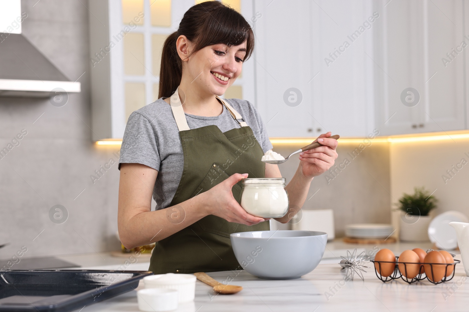 Photo of Happy young housewife adding flour into bowl at white marble table in kitchen. Cooking process