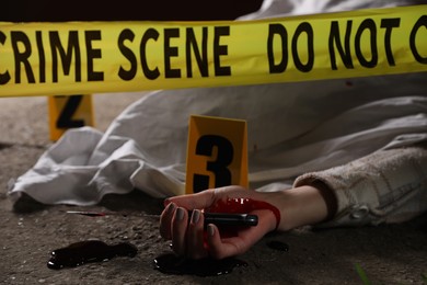 Crime scene with dead woman's body, bloody knife, markers and yellow tape outdoors, closeup