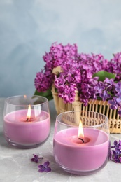 Burning wax candles in glass holders and lilac flowers on grey table, space for text