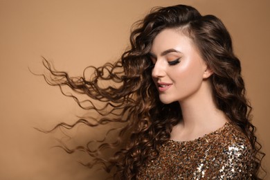 Photo of Beautiful young woman with long curly brown hair in golden sequin dress on beige background, space for text