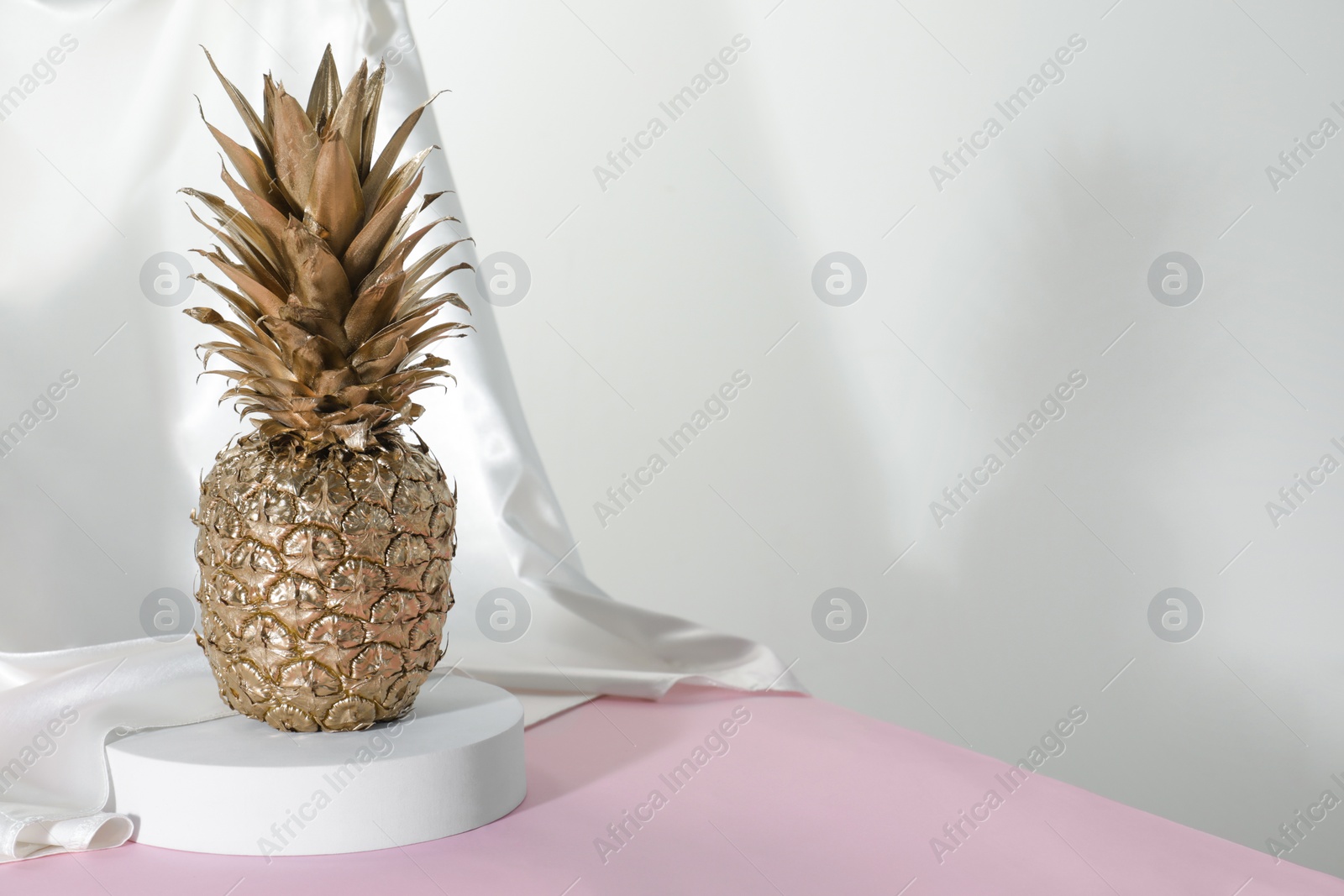 Photo of Golden pineapple, round shaped podium and silk fabric on pink table against white background. Space for text