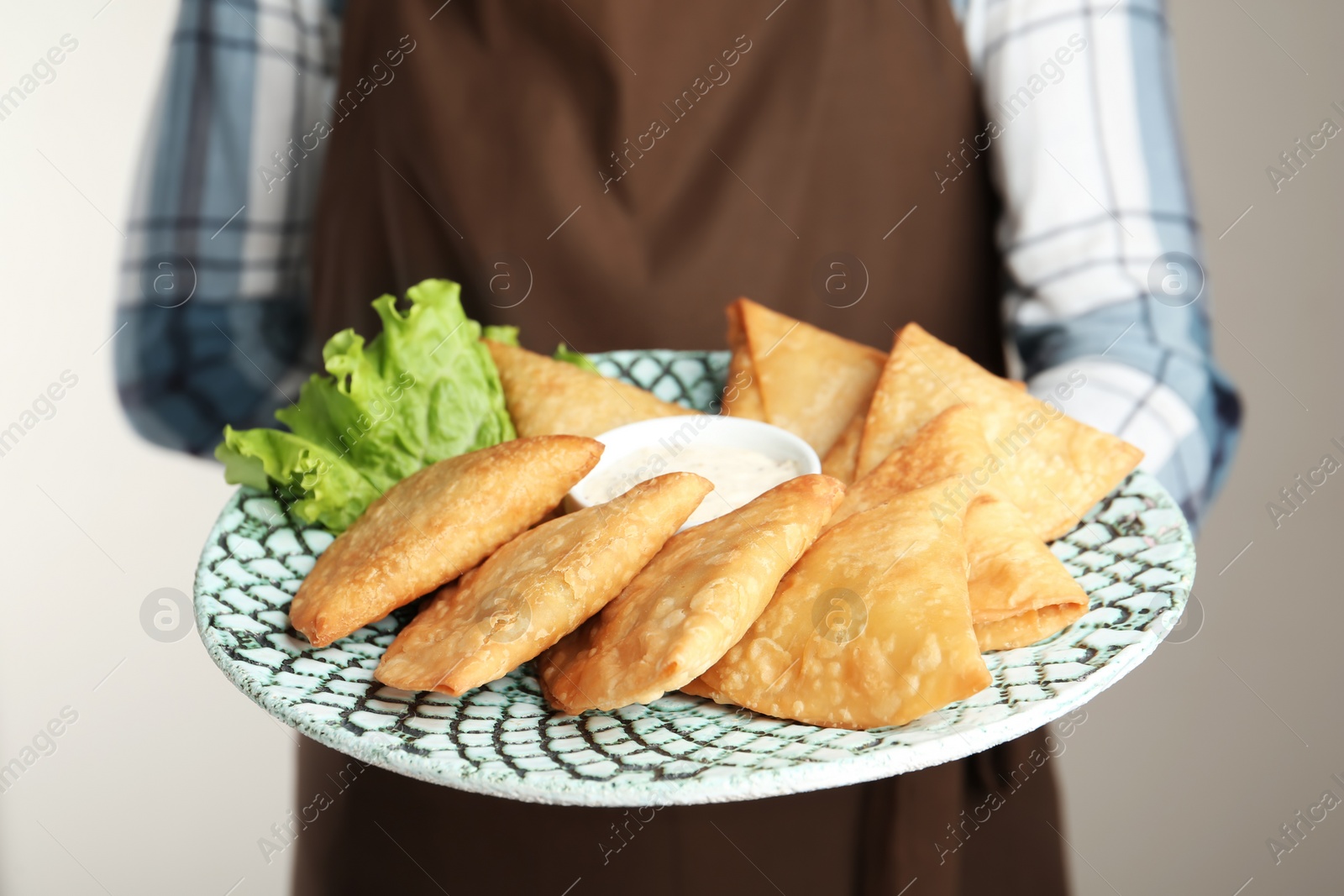 Photo of Woman holding plate with delicious samosas and sauce on light grey background, closeup