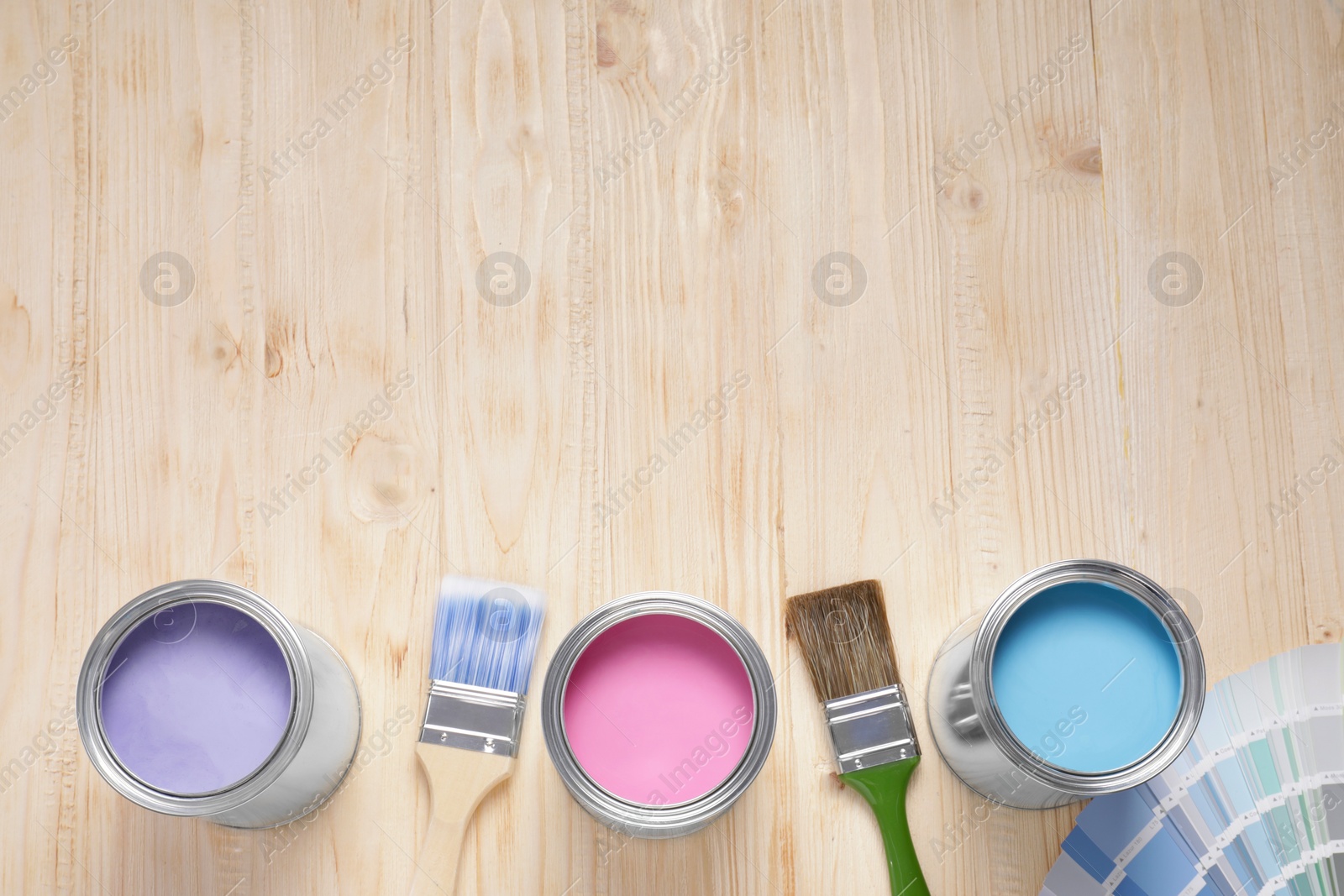 Photo of Cans of lilac, pink and light blue paints, palette with brushes on wooden table, flat lay. Space for text