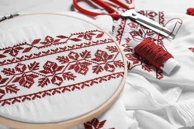 Photo of Shirt with red embroidery design in hoop, scissors and thread on table, closeup. National Ukrainian clothes