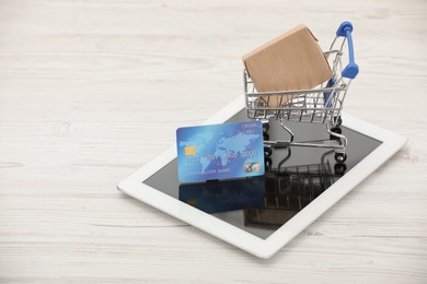 Photo of Online payment concept. Small shopping cart with bank card, box and tablet on white table, space for text