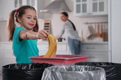 Photo of Little girl throwing banana peel into trash bin in kitchen. Separate waste collection