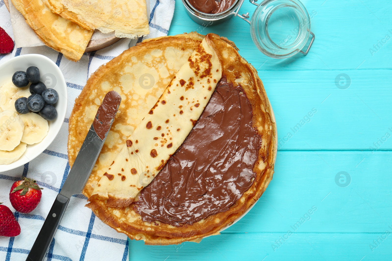 Photo of Tasty crepes with chocolate paste and berries served on turquoise wooden table, flat lay