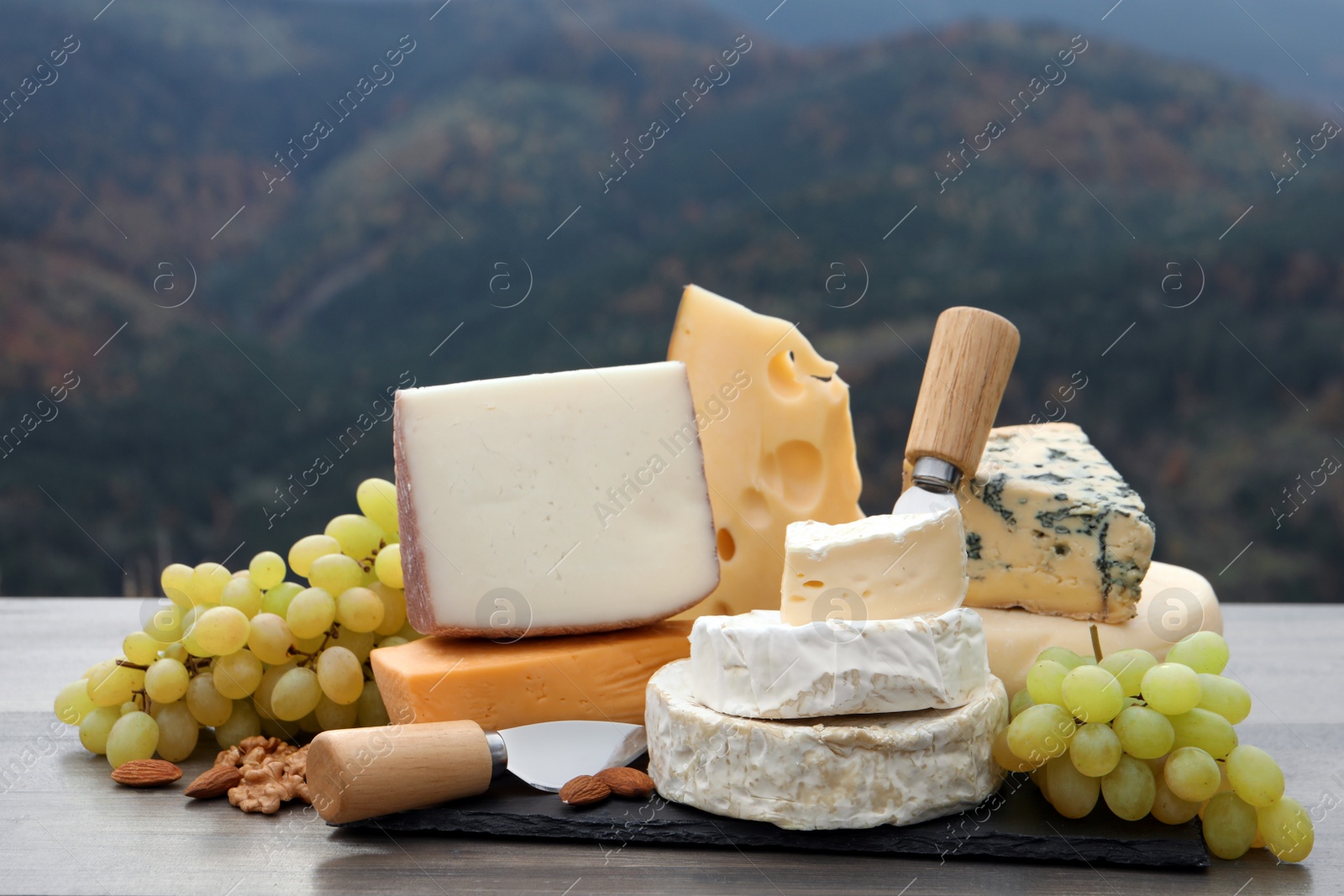 Photo of Different types of delicious cheeses and snacks on wooden table against mountain landscape