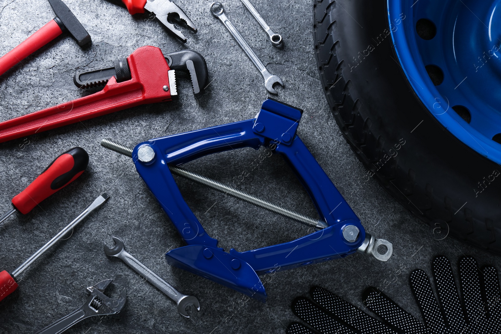 Photo of Car wheel, scissor jack, gloves and different tools on grey stone surface, flat lay