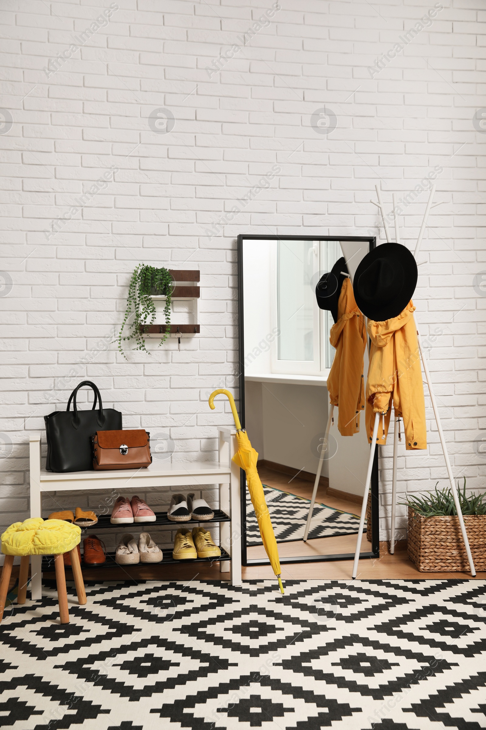 Photo of Stylish hallway interior with coat rack, shoe storage bench and mirror near white brick wall