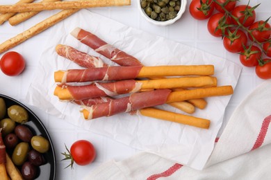 Delicious grissini sticks with prosciutto and snacks on white table, flat lay