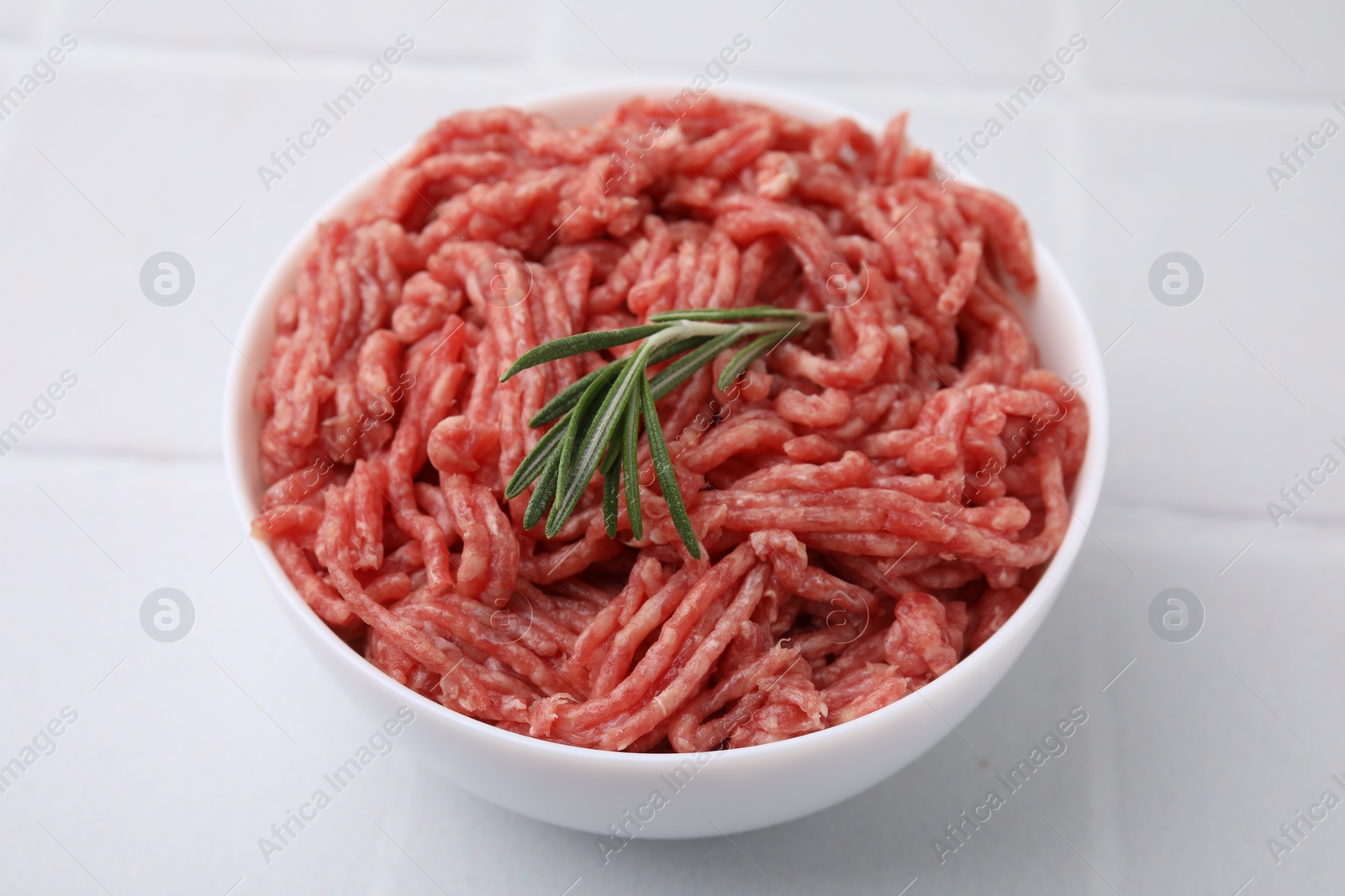 Photo of Fresh raw ground meat and rosemary in bowl on white tiled table, closeup