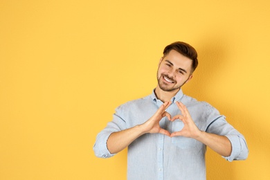 Portrait of young man making heart with his hands on color background, space for text