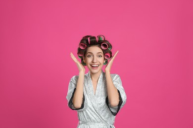 Photo of Emotional young woman in silk bathrobe with hair curlers on pink background