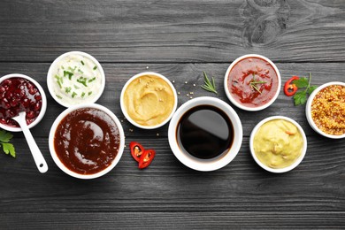 Photo of Different tasty sauces in bowls, parsley, chili pepper and rosemary on black wooden table, flat lay