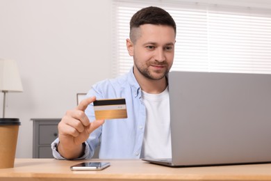 Photo of Handsome man with credit card using laptop for online shopping at wooden table in room