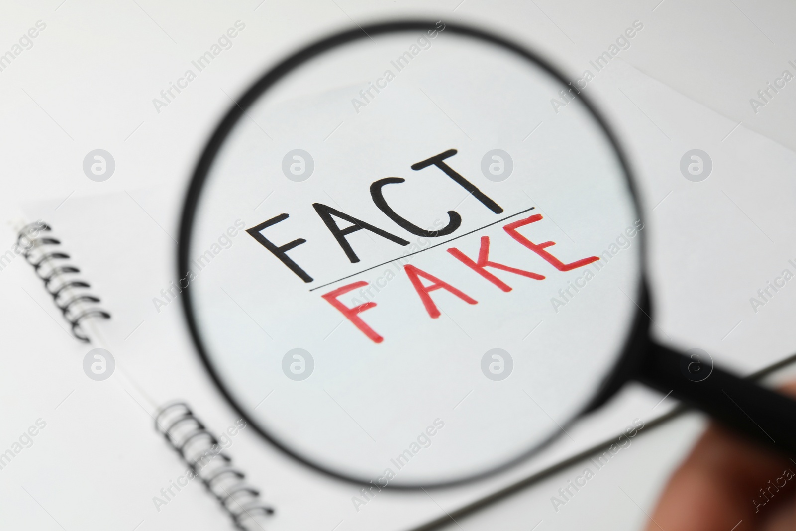 Photo of Woman holding magnifying glass over notebook with words Fact and Fake on white background, closeup