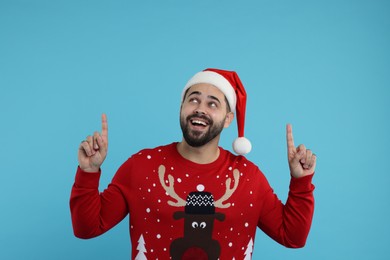 Photo of Happy young man in Christmas sweater and Santa hat pointing at something on light blue background