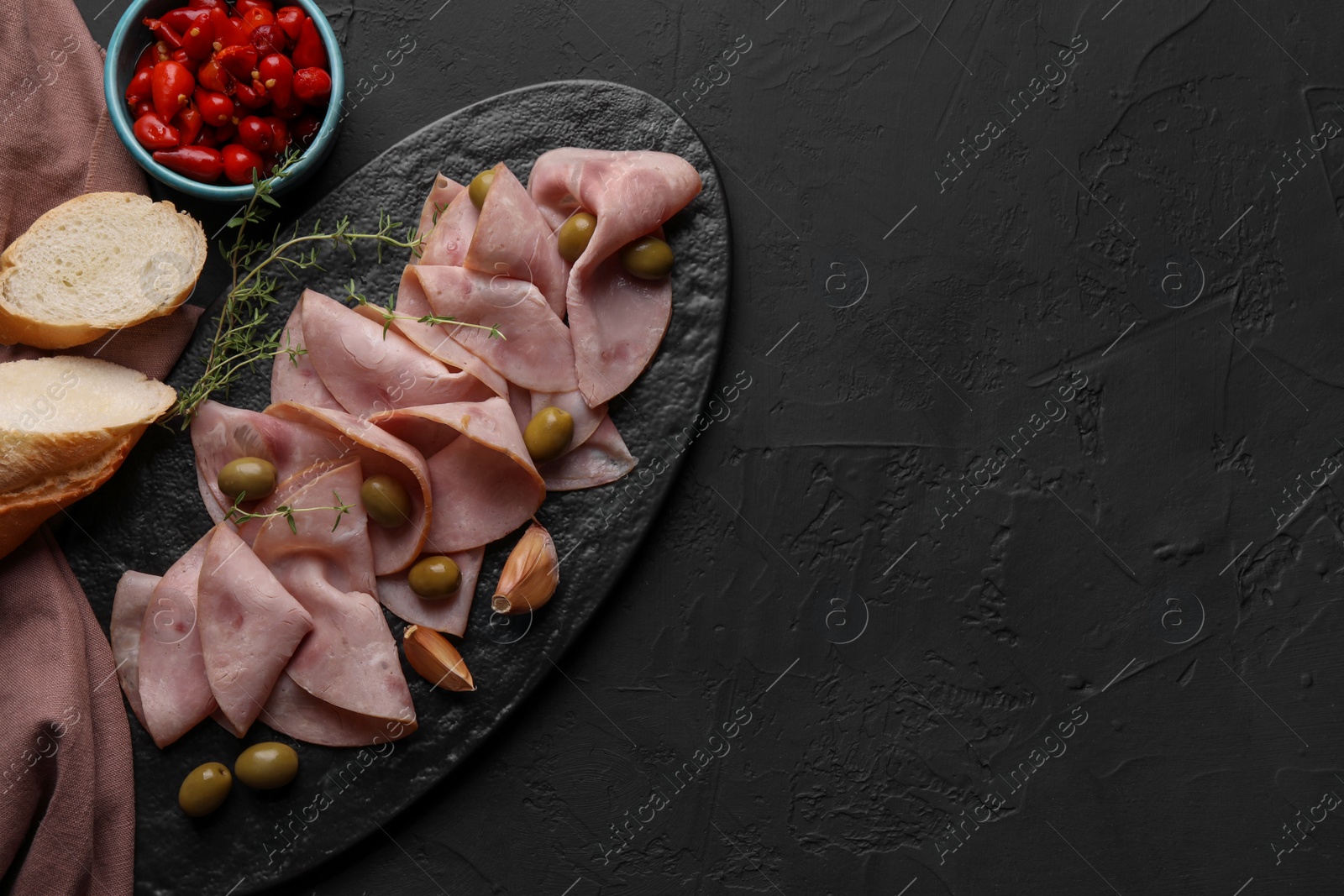 Photo of Tasty ham with olives, garlic, bread and pickled peppers on black textured table, flat lay