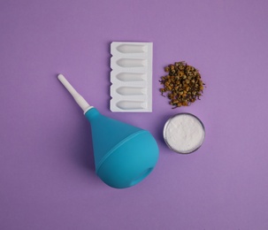 Enema, suppositories, dry chamomile and bowl of salt on violet background, flat lay