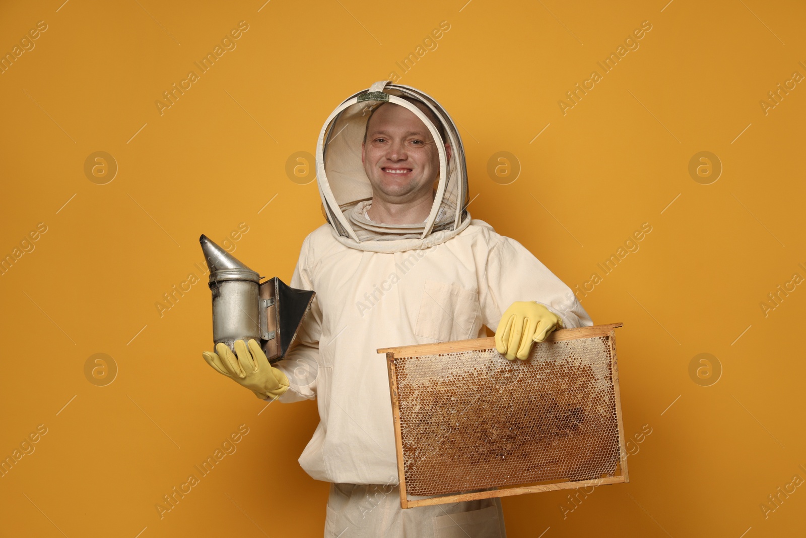 Photo of Beekeeper in uniform holding smokepot and hive frame with honeycomb on yellow background