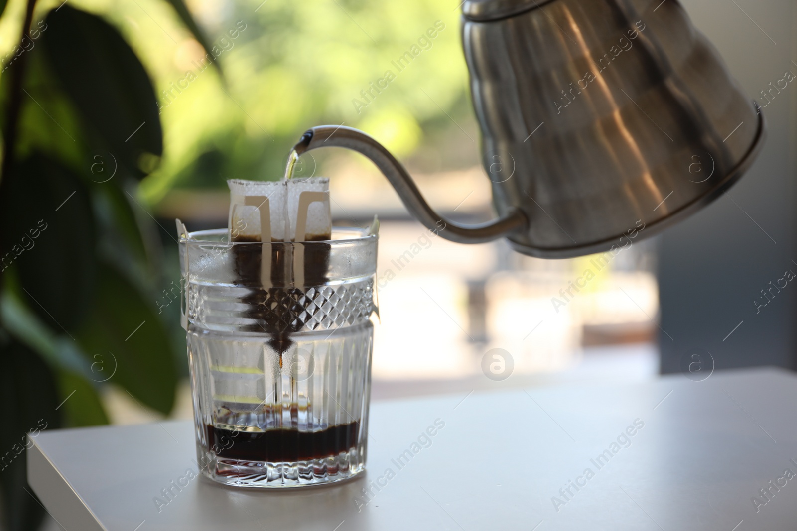 Photo of Pouring hot water into glass with drip coffee bag from kettle on white table