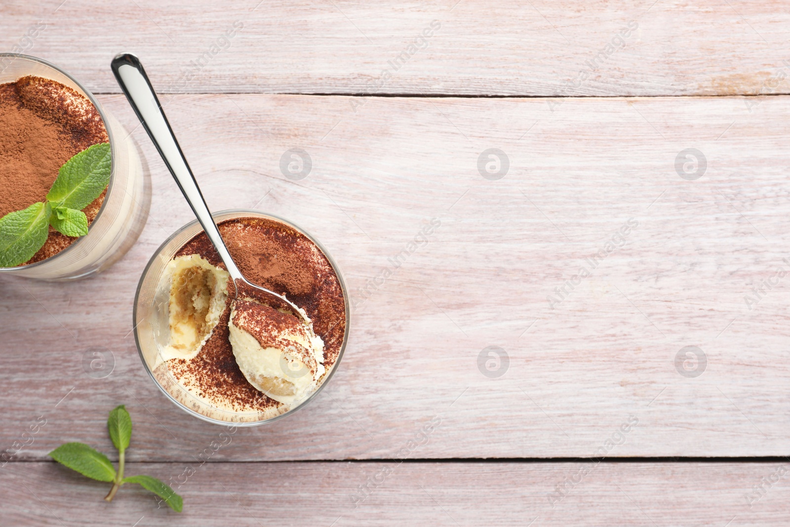 Photo of Delicious tiramisu in glasses, mint leaves and spoon on wooden table, top view. Space for text