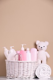 Photo of Wicker basket with baby cosmetic products, bath accessories and knitted toy bear on white table against beige background