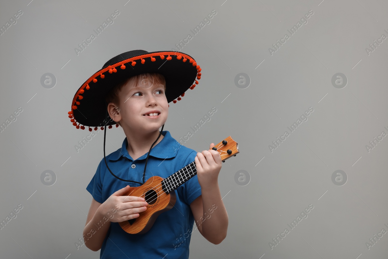 Photo of Cute boy in Mexican sombrero hat playing ukulele on grey background, space for text