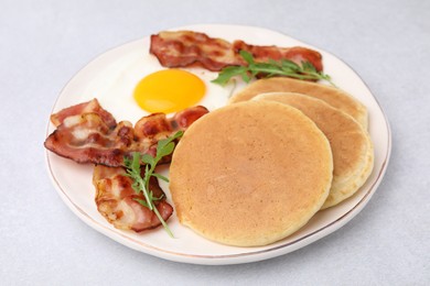 Photo of Plate with tasty pancakes, fried egg, arugula and bacon on light grey textured table, closeup