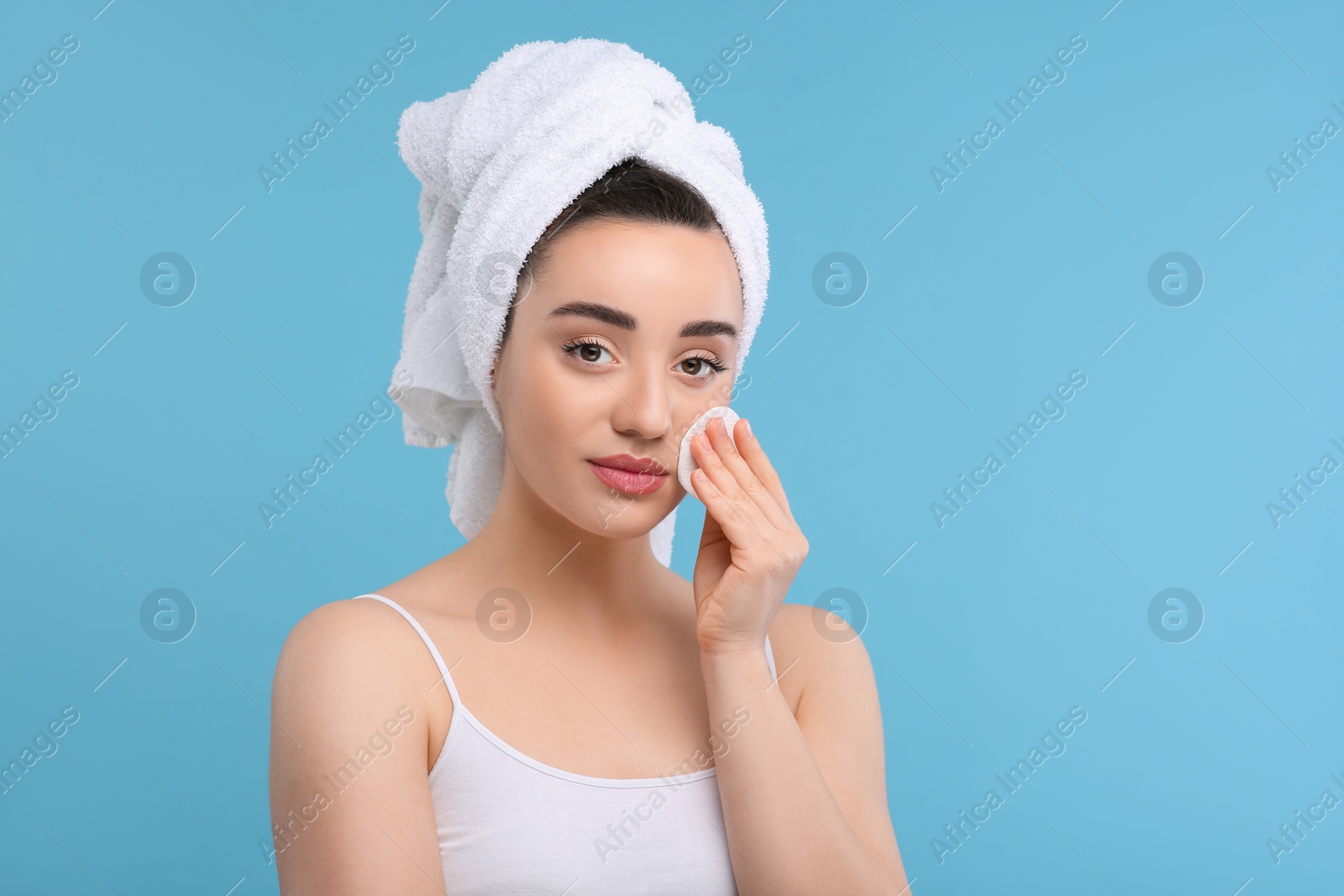 Photo of Beautiful woman in terry towel removing makeup with cotton pad on light blue background, space for text