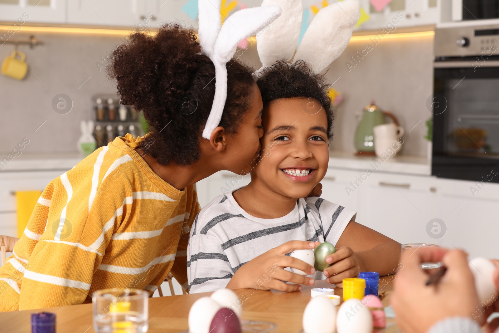 Photo of Happy African American mother kissing her cute son while painting Easter eggs at table in kitchen