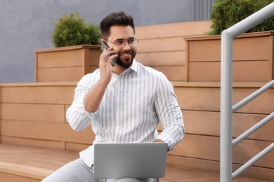 Photo of Handsome young man talking on smartphone while using laptop on bench outdoors
