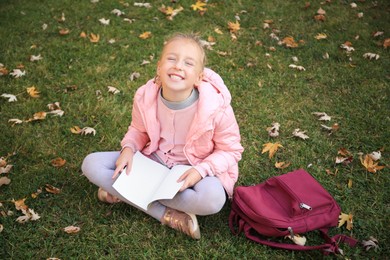 Photo of Cute little girl with copybook and backpack on green grass outdoors