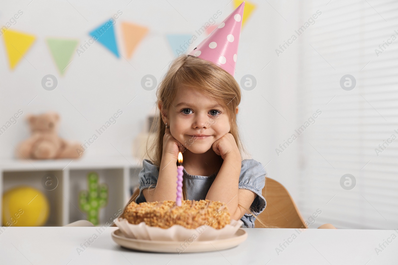 Photo of Cute girl in party hat with birthday cake at table indoors