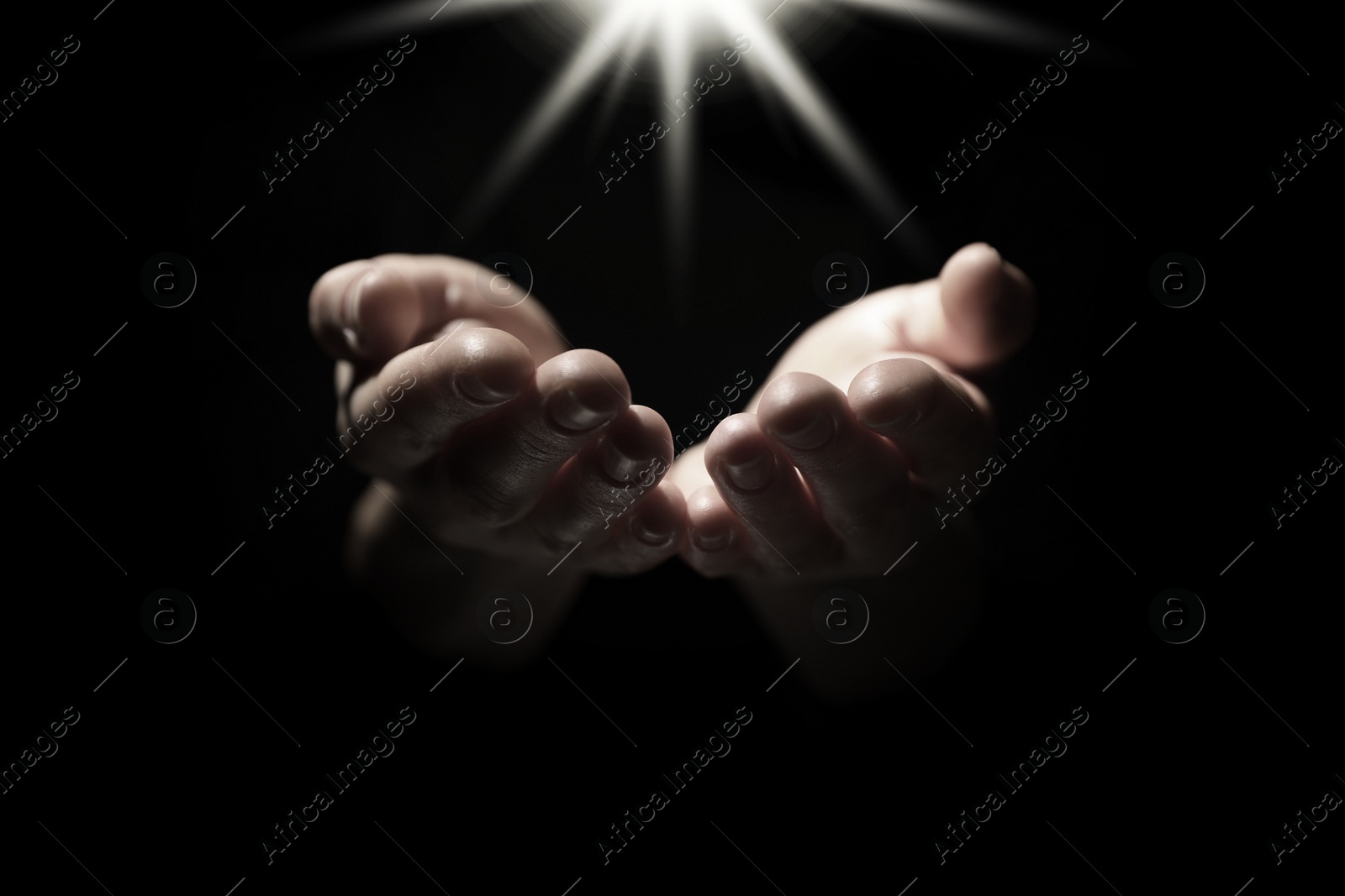 Image of Christian woman holding hands under holy light in darkness, closeup. Prayer and belief