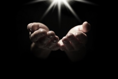 Image of Christian woman holding hands under holy light in darkness, closeup. Prayer and belief
