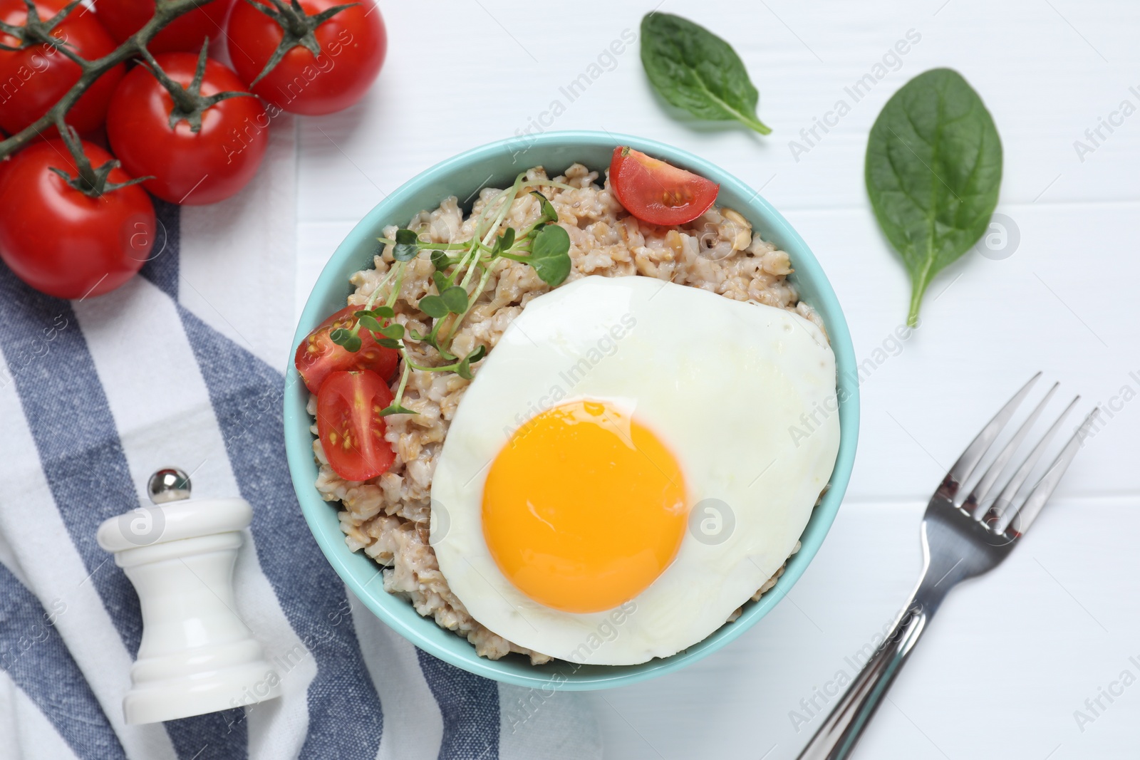 Photo of Tasty boiled oatmeal with fried egg, tomato and microgreens served on white wooden table, flat lay