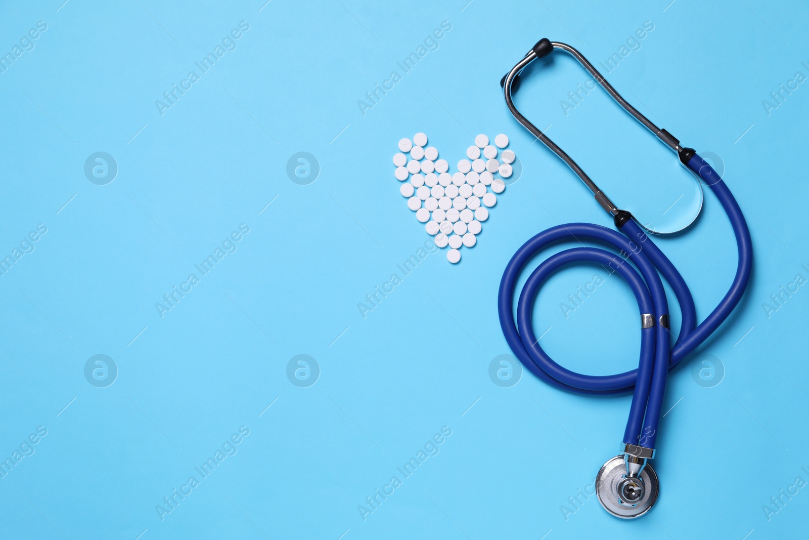 Photo of Heart shape made of pills near stethoscope on light blue background, flat lay. Space for text