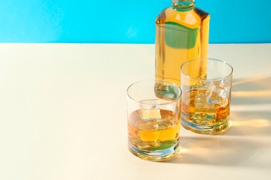 Whiskey with ice cubes in glasses and bottle on white table, space for text