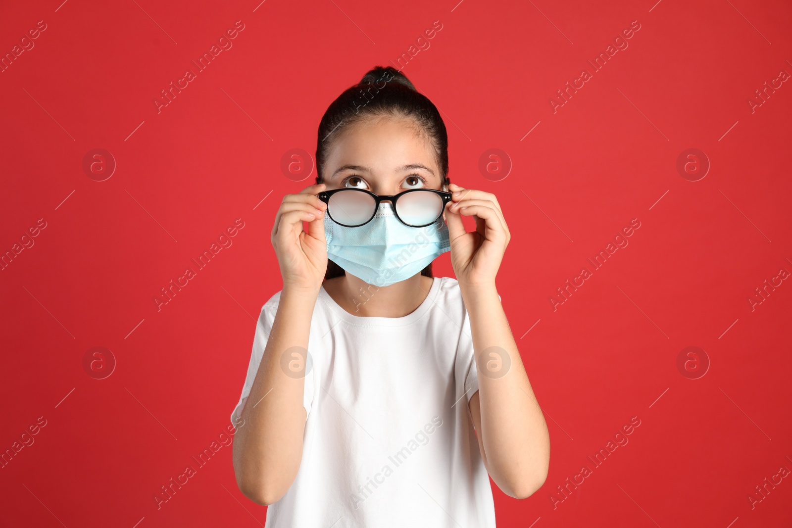 Photo of Little girl wiping foggy glasses caused by wearing medical face mask on red background. Protective measure during coronavirus pandemic