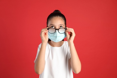 Little girl wiping foggy glasses caused by wearing medical face mask on red background. Protective measure during coronavirus pandemic