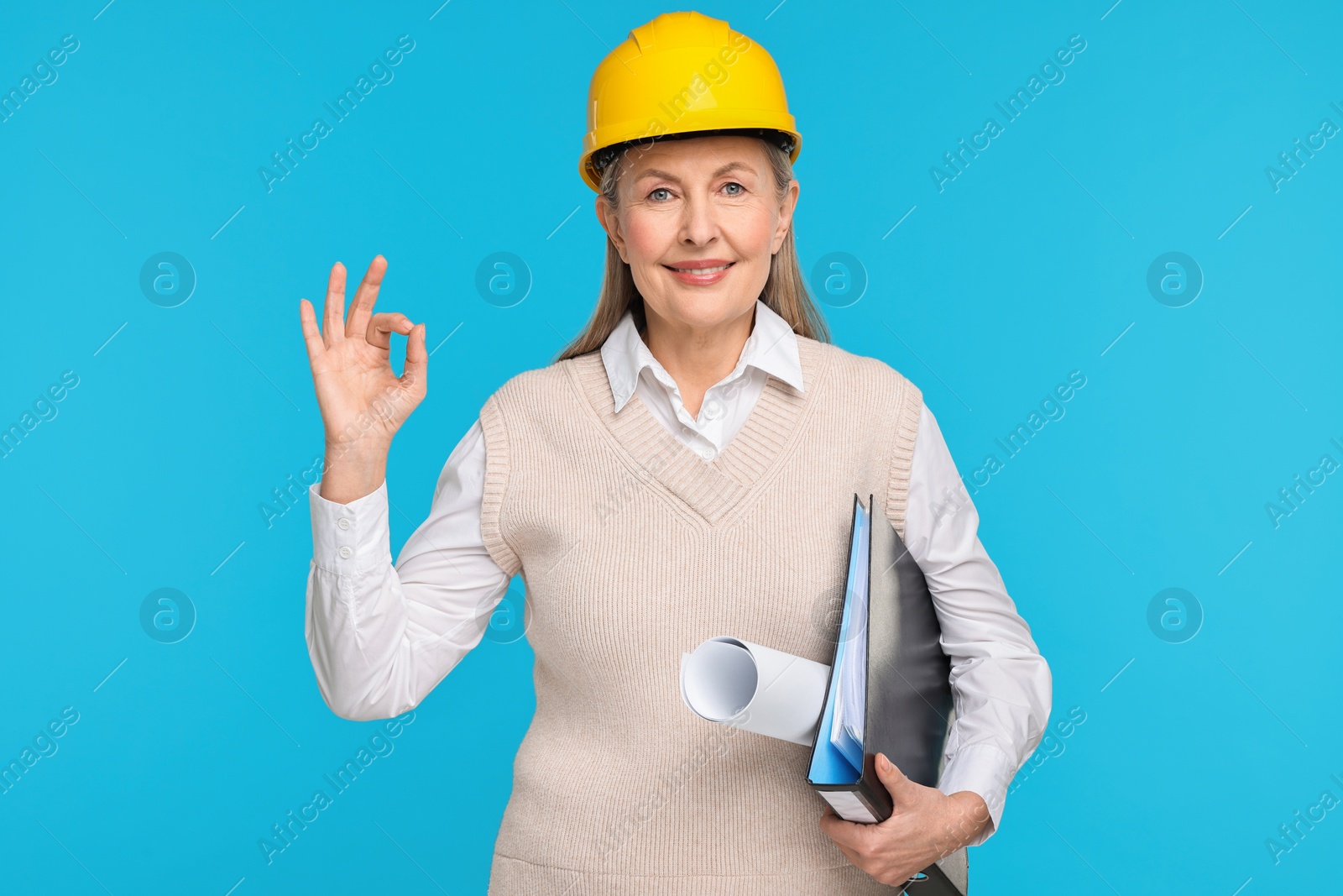 Photo of Architect in hard hat with draft and folder showing ok gesture on light blue background