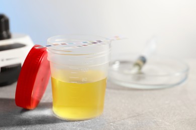 Photo of Container with urine sample for analysis and test strips on grey table in laboratory, closeup. Space for text