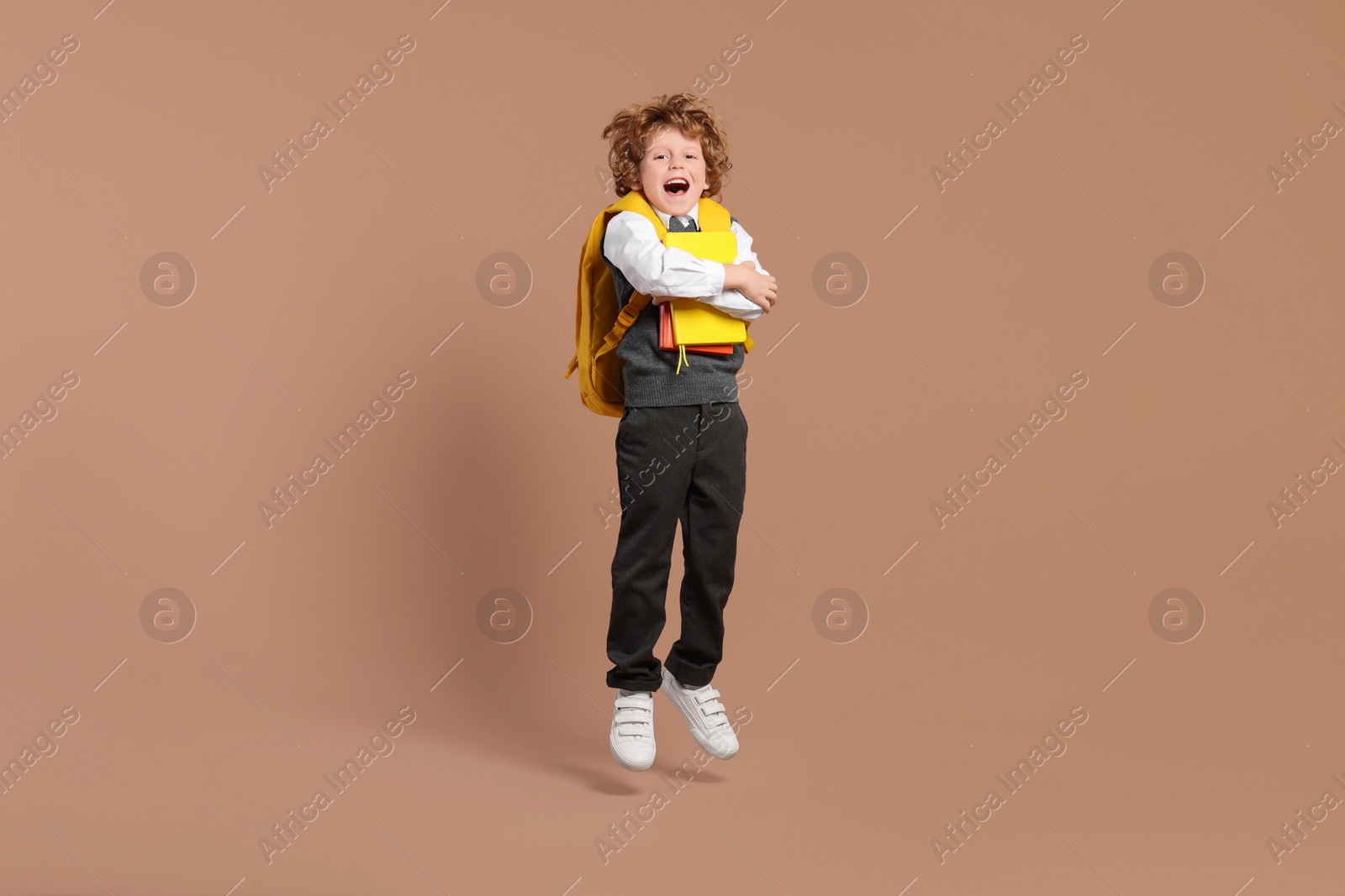 Photo of Happy schoolboy with backpack and books jumping on brown background